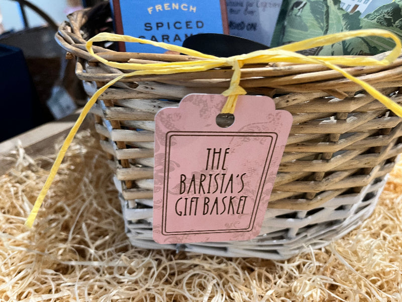 Wicker gift basket with a pink tag labeled 'The Barista's Gift Basket' on a bed of straw.