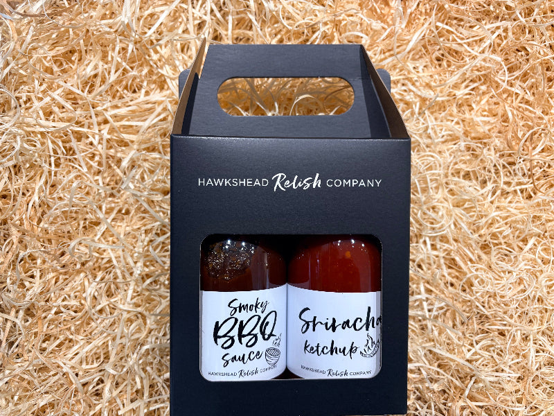 Two bottles of sauce in a black box with 'Hawkshead Relish Company' branding on a straw background.